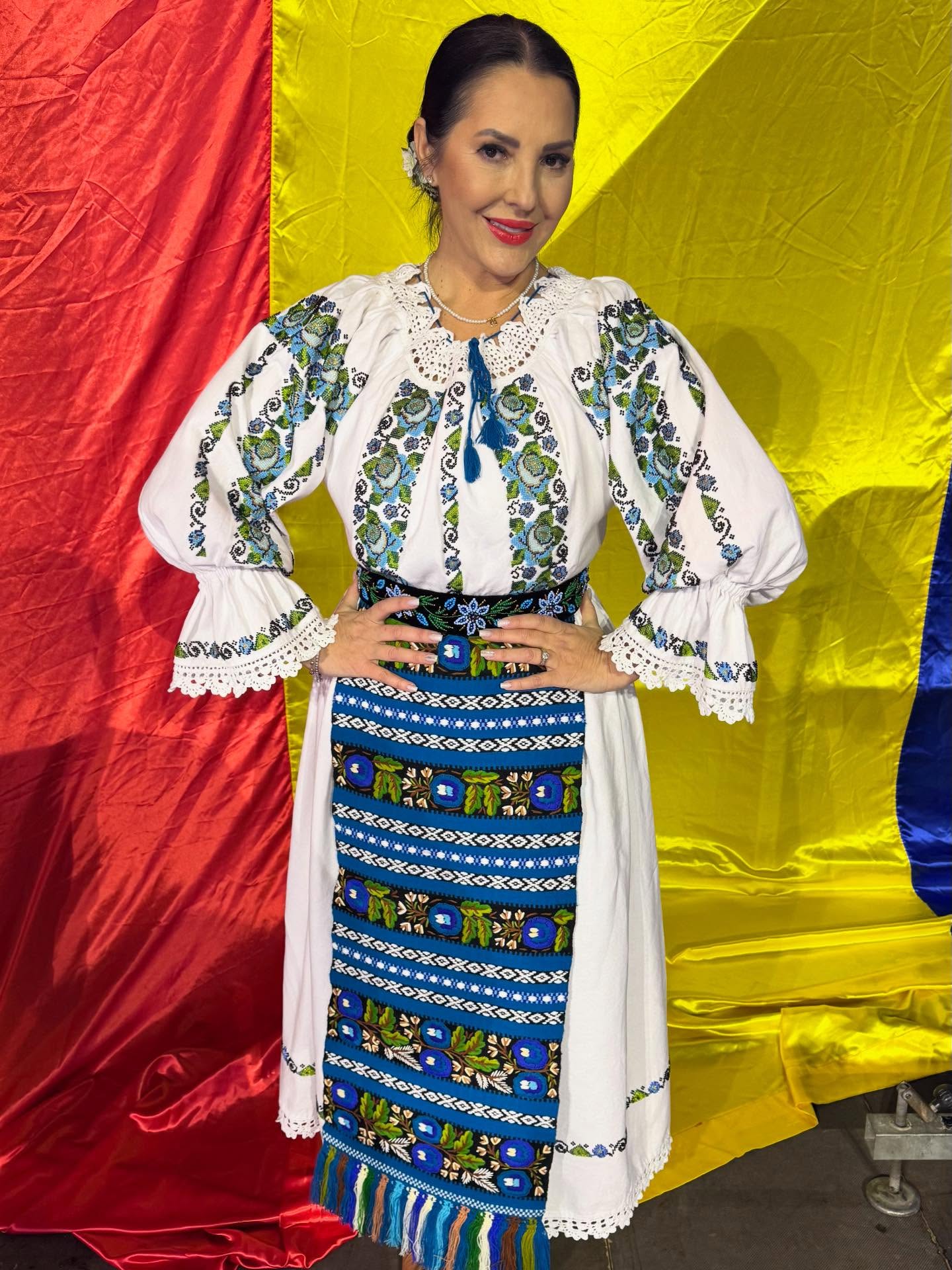 Angela Rusu z&acirc;mbitoare, cu m&acirc;inile &icirc;n șold, &icirc;n costum popular alb cu albastru și verde, cu părul prins &icirc;ntr-un coc la spate