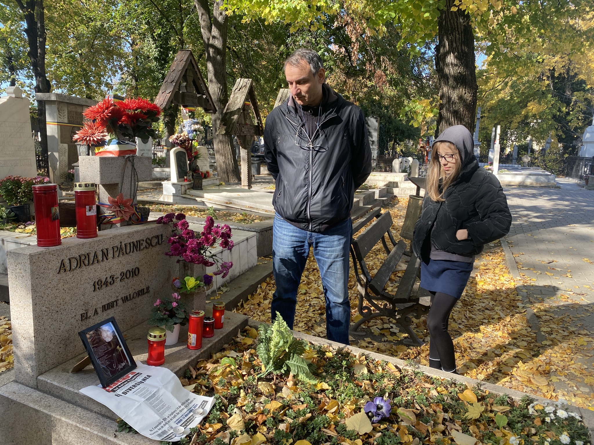 Andrei Păunescu &icirc;ntr-o poziție solemnă, cu m&acirc;inile la spate și privirea &icirc;n păm&acirc;nt, la morm&acirc;ntul lui Adrian Păunescu, alături de fiica lui cea mică, Andreea Păunescu