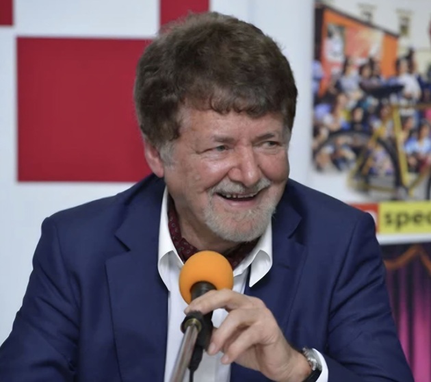 Compozitorul&nbsp;Jolt&nbsp;Kerestely z&acirc;mbitor, &icirc;ntr-un costum albastru &icirc;nchis, cu cămașă albă, z&acirc;mbitor, st&acirc;nd pe scaun și țin&acirc;nd &icirc;n m&acirc;na st&acirc;ngă un microfon