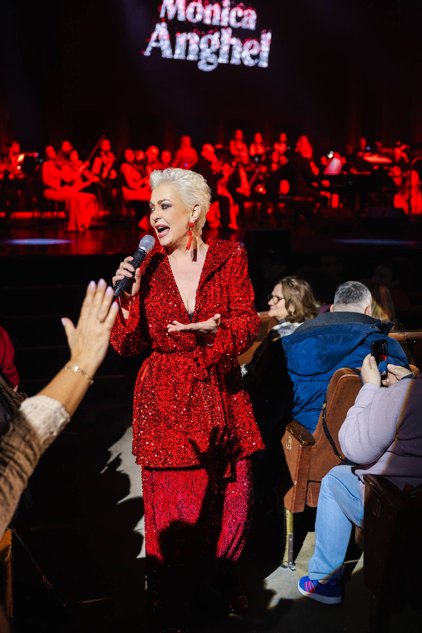 Monica Anghel &icirc;ntr-un costum roșu cu paiete, c&acirc;nt&acirc;nd &icirc;n mijlocul publicului său, printre scaunele de la Sala Palatului