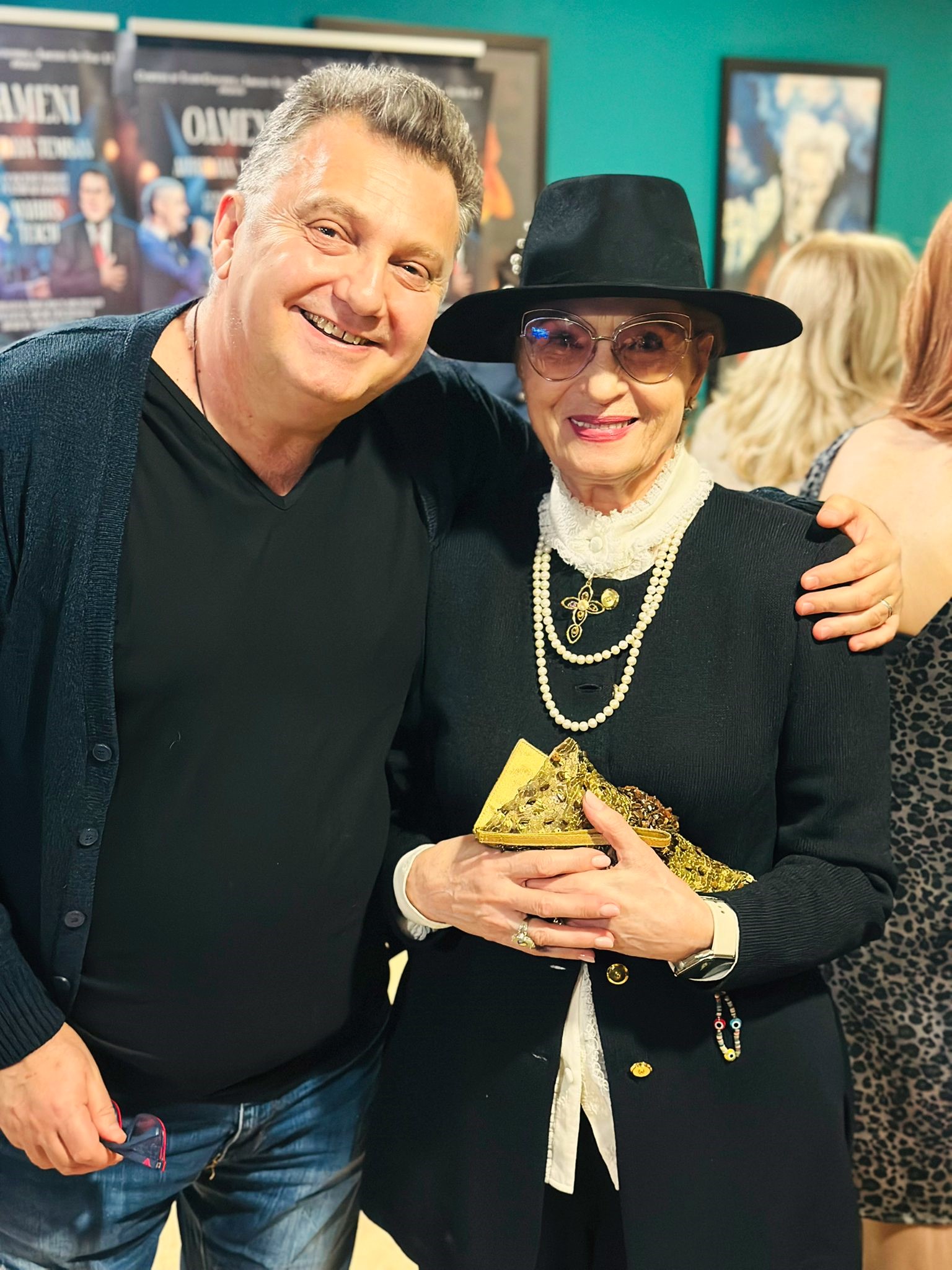 Compozitorul Mihai Alexandru z&acirc;mbitor, str&acirc;ng&acirc;nd-o &icirc;n brațe pe Angela Similea, &icirc;mbrăcată &icirc;n negru și alb, cu pălărie pe cap și ochelari de mari dimensiuni la ochi