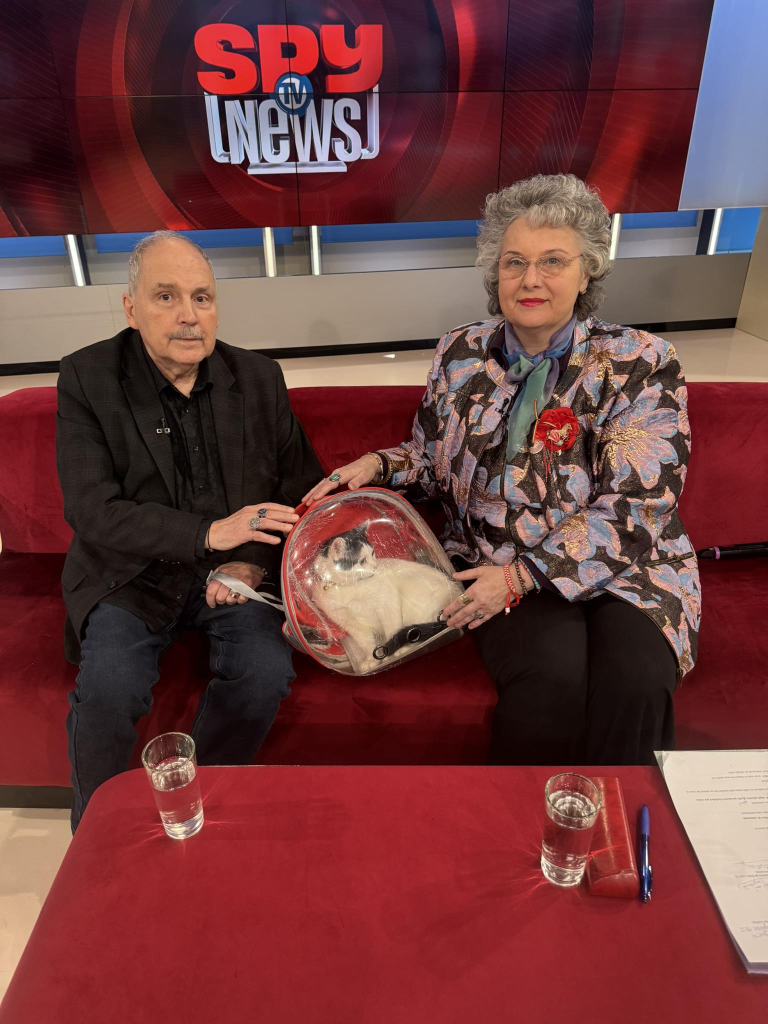 Eugen Cristea, &icirc;mbrăcat &icirc;n negru, st&acirc;nd pe canapea &icirc;n platoul Spy News Tv, cu pisica pe care a primit-o cadou și &icirc;n care susține că s-a re&icirc;ncarnat soția lui
