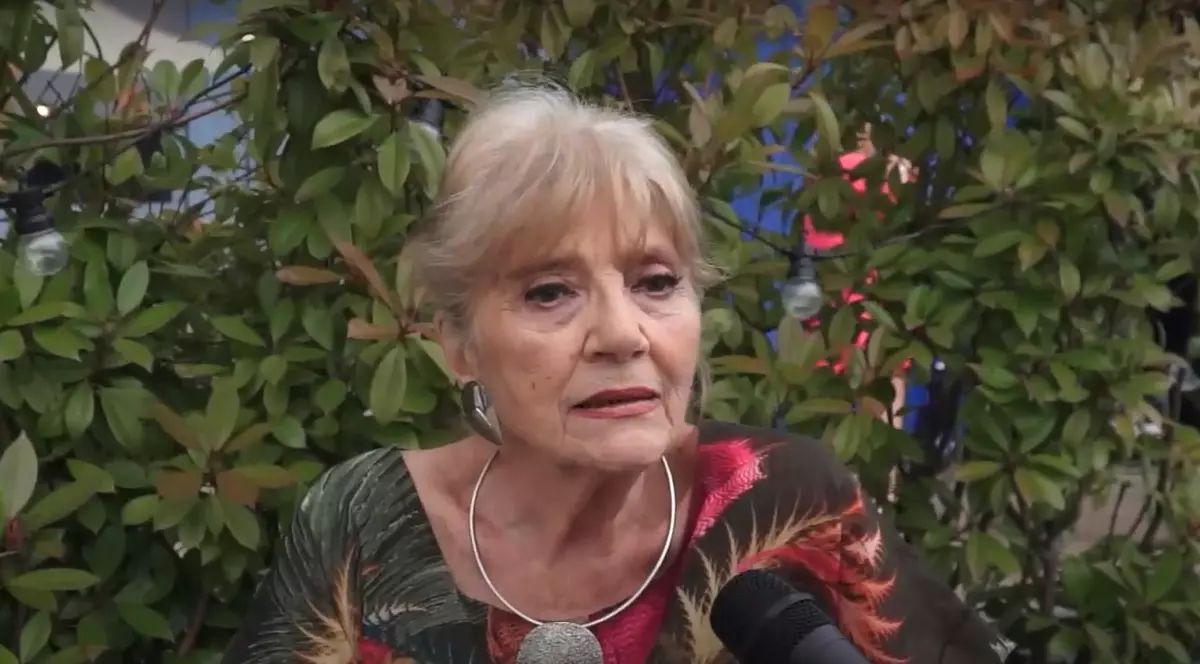 Regretata actriță Cristina Deleanu, &icirc;mbrăcată &icirc;ntr-o rochie neagră cu imprimeu floral, st&acirc;nd l&acirc;ngă un gard viu, &icirc;n timp ce acorda un interviu