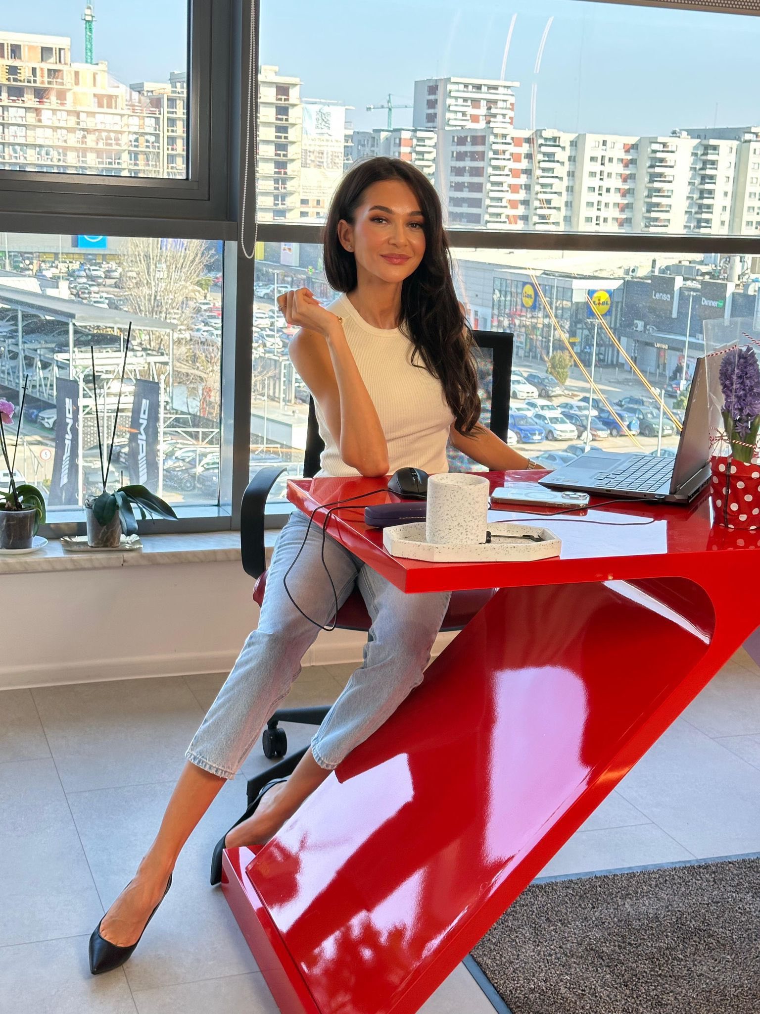 Ruxandra Luca &icirc;ntr-un birou cu geamuri mari, st&acirc;nd &icirc;n fața laptopului, &icirc;mbrăcată &icirc;ntr-un top alb, cu blugi deschiși la culoare, purt&acirc;nd &icirc;n picioare pantofi stiletto