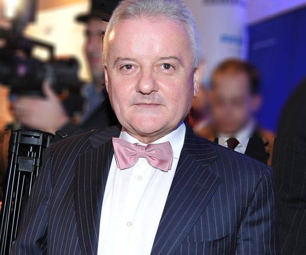 Irinel Columbeanu &icirc;ntr-un costum bleumarin cu dungi albe subțiri, cămașă albă și papion roz, la un eveniment de seară