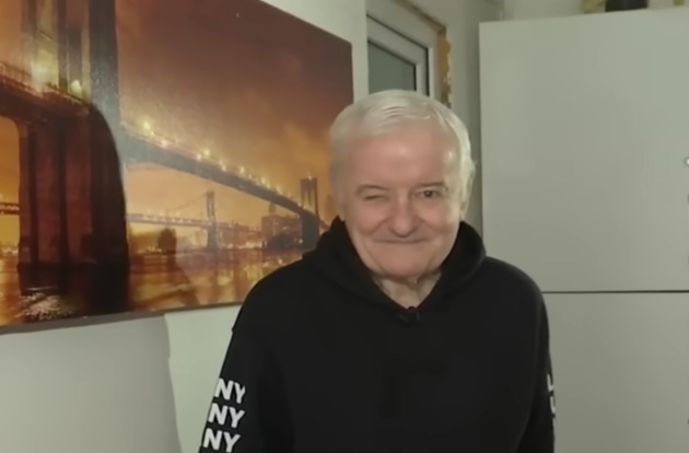 Irinel Columbeanu &icirc;ntr-un hanorac negru, cu scris alb pe m&acirc;neci, st&acirc;nd &icirc;ntr-o cameră a azilului din Ghermănești &icirc;n care locuiește