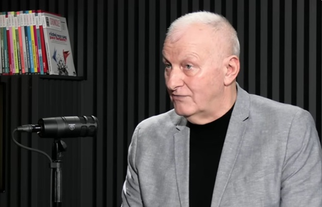 Generalul &icirc;n rezervă Virgil&nbsp;Bălăceanu, st&acirc;nd la masă și vorbind la microfon, &icirc;n cadrul unui podcast, &icirc;n timp ce purta un tricou negru și un sacou gri