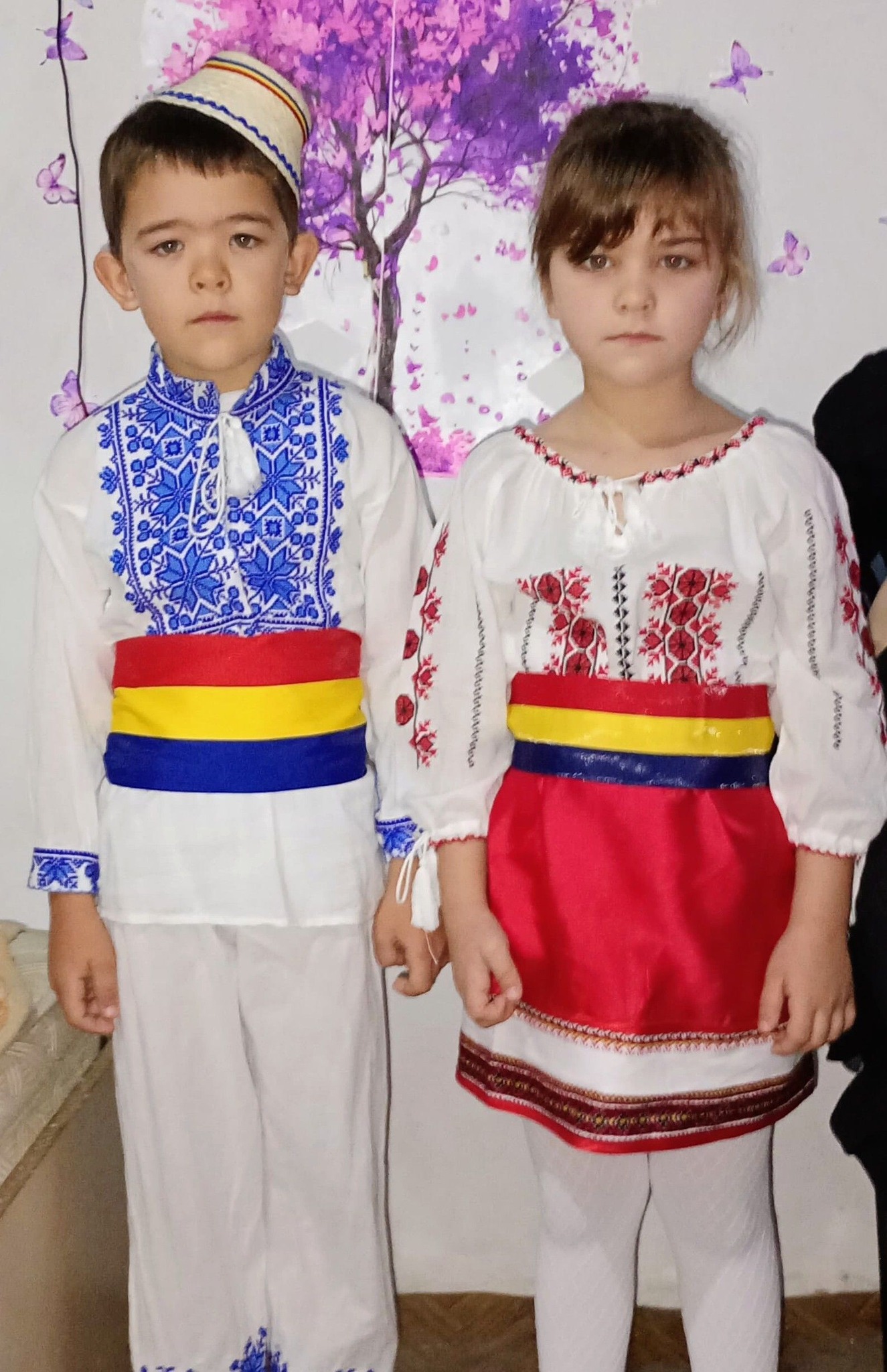 Irina și Robert Neacșu &icirc;mbrăcați &icirc;n costum popular, la serbarea de la grădiniță, st&acirc;nd serioși la poză, l&acirc;ngă un perete cu desene coloarate