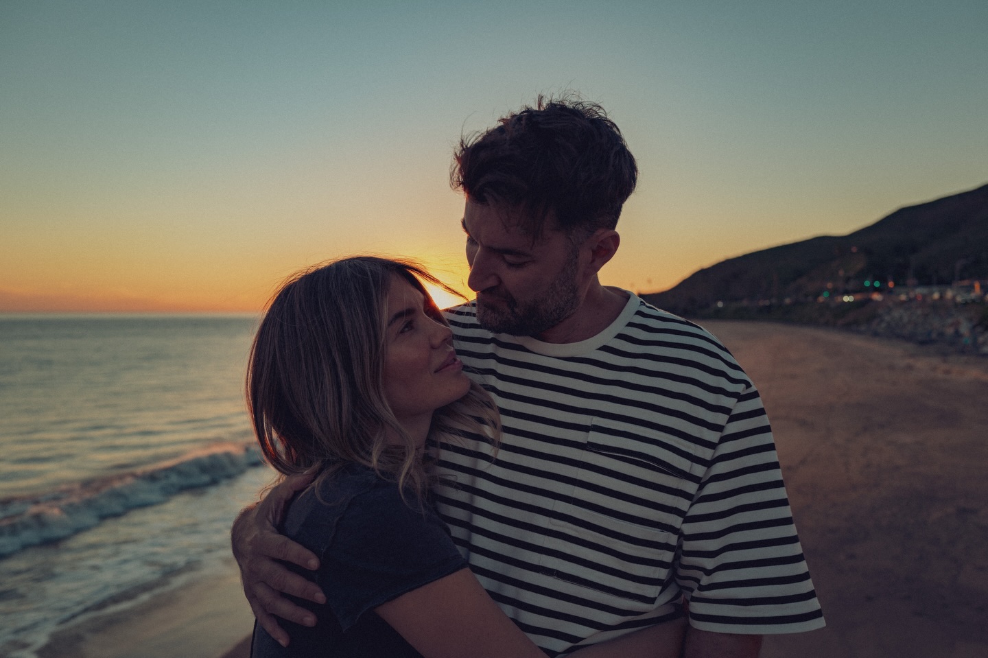 Gina Pistol și Smiley, pe plajă, la apus, țin&acirc;ndu-se &icirc;n brațe și privindu-se &icirc;n ochi