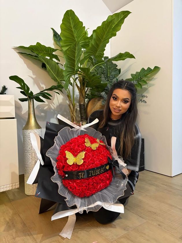 Adriana Simionescu brunetă, îmbrăcată în negru, stând pe podea cu picioarele încrucișate, ținând în brațe un buchet mare de trandafiri, care are pe mijloc o panglică pe care scrie „Te iubesc”