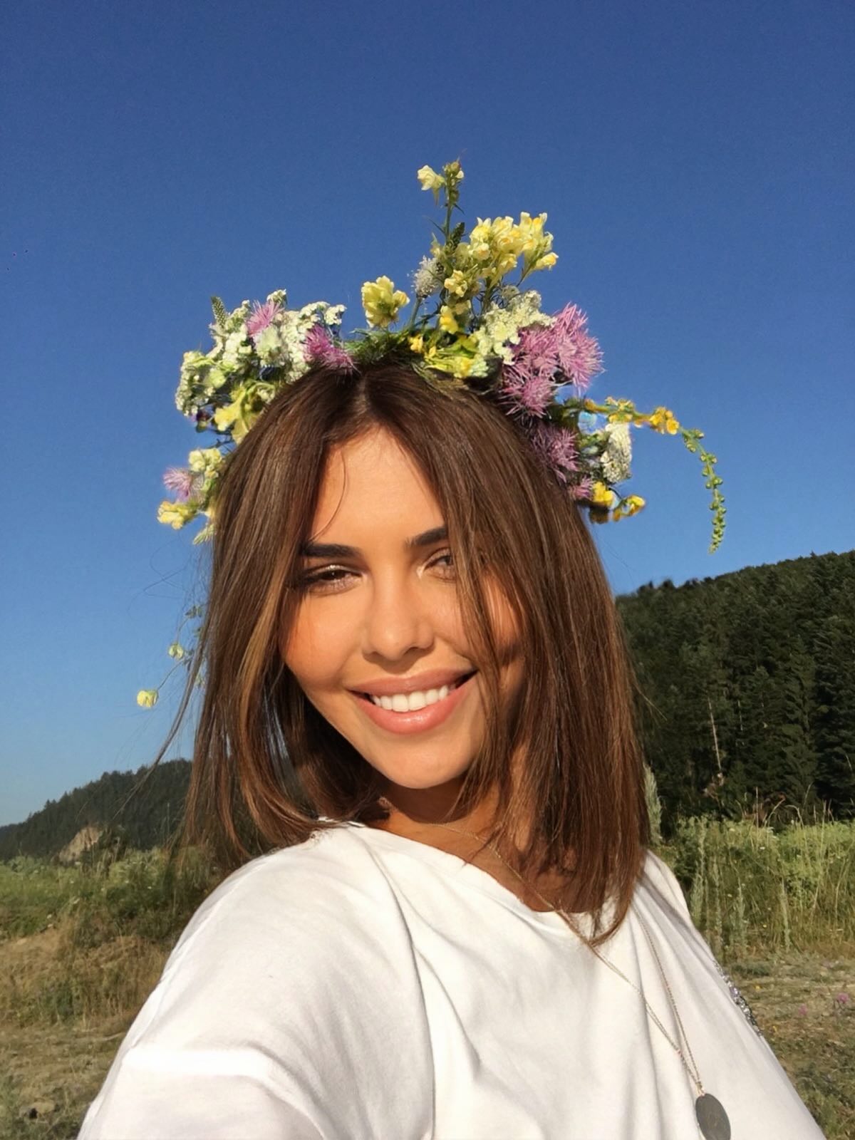 Andreea Popescu z&acirc;mbitoare, &icirc;mbrăcată &icirc;n alb, făc&acirc;ndu-și un selfie &icirc;n timp ce poartă pe cap o coroană din flori de c&acirc;mp