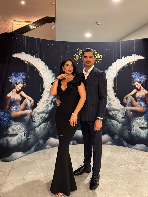 Deea Maxer și logodnicul ei, Constantin Cataramă, la un eveniment de seară, &icirc;mbrăcați am&acirc;ndoi &icirc;n negru, &icirc;n ținute elegante, st&acirc;nd &icirc;mbrățișați &icirc;n fața unui panou foto