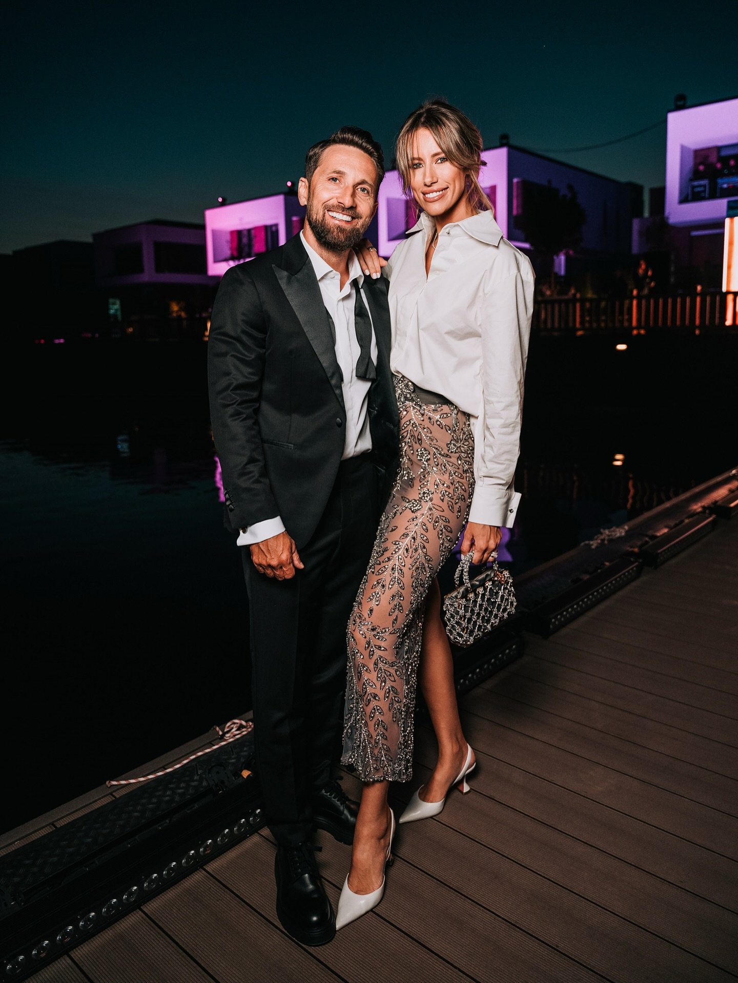 Gabriela&nbsp;Prisăcariu și soțul ei, Dani Oțil, z&acirc;mbitori pe un ponton, &icirc;mbrăcați elegant, la un eveniment de seară, el &icirc;n costum negru și cămașă albă, iar ea &icirc;n fustă bej cu paiete și cămașă albă