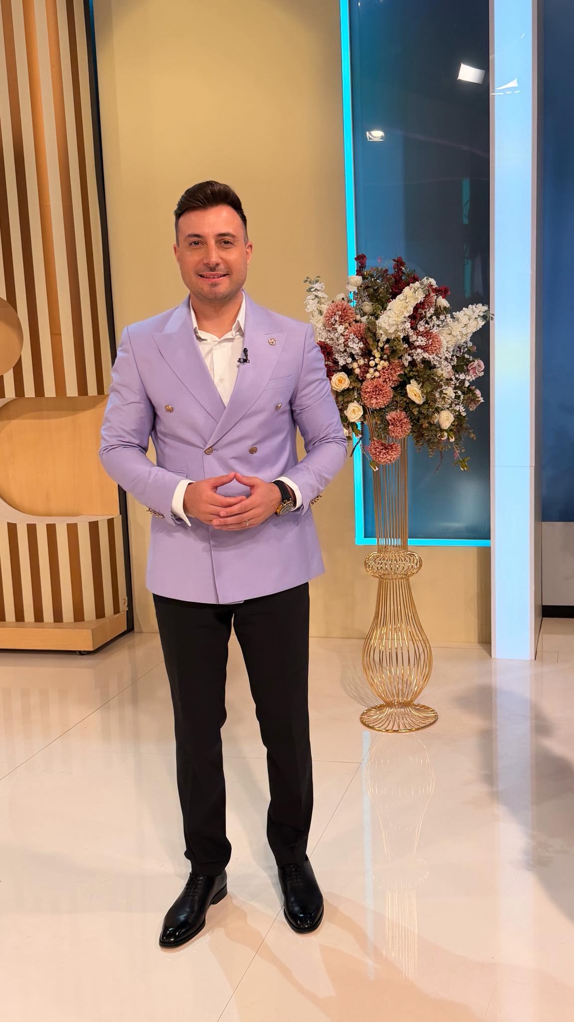 Valentin Sanfira &icirc;n platoul unei emisiuni TV, &icirc;mbrăcat &icirc;n pantaloni negri, sacou lila și cămașă albă, țin&acirc;ndu-și m&acirc;inile &icirc;mpreunate cu verigheta la vedere