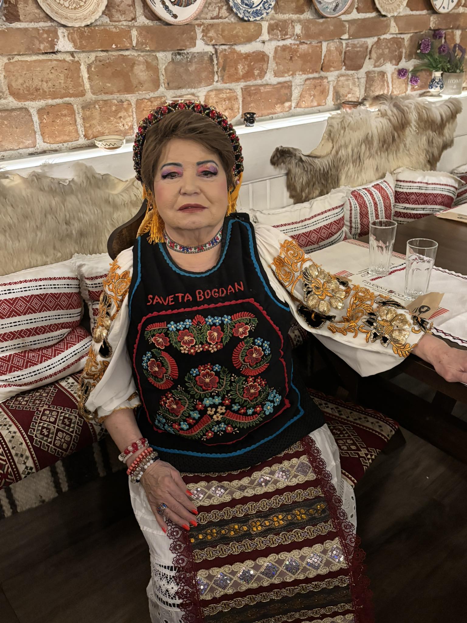 Saveta&nbsp;Bogdan &icirc;n costum popular peste care are o vestă cu motive tradiționale, pe care scrie numele ei, &icirc;n timp ce stă pe scaun, la masa unui restaurant decorat &icirc;n stil rustic