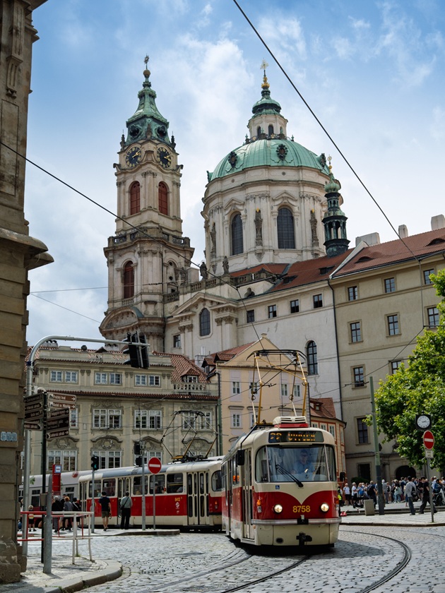 Tramvai roșu, în mers, pe o stradă din Praga, chiar lângă Biserica Sfântul Nicolae, în timp ce troruarul este plin de oameni