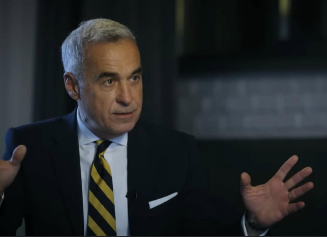 Călin Georgescu fotografiat &icirc;n timp ce vorbește și gesticulează, &icirc;mbrăcat &icirc;n costum negru, cămașă albă și cravată &icirc;n dungi.