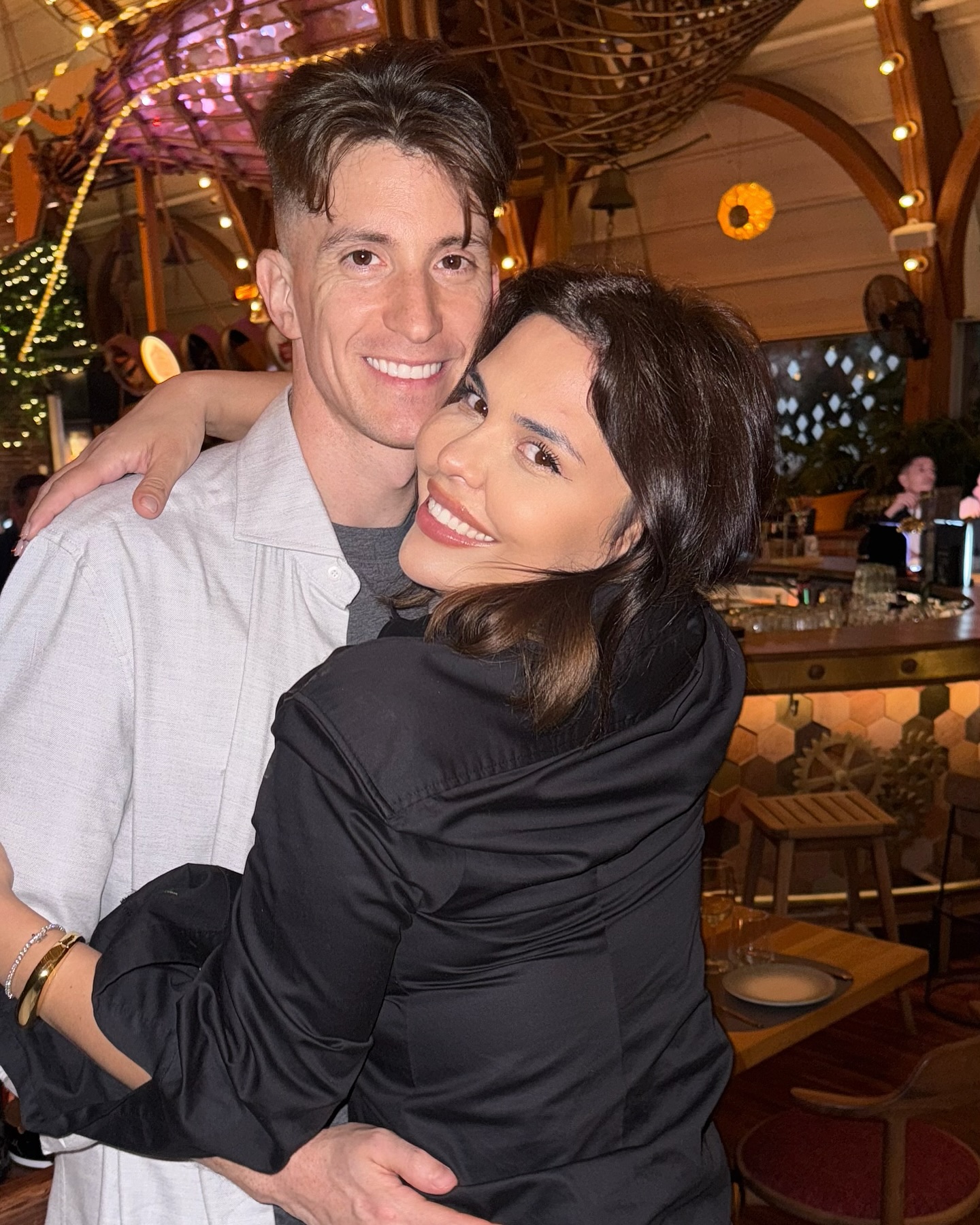 Andreea Popescu z&acirc;mbitoare, &icirc;mbrățiș&acirc;ndu-l str&acirc;ns pe Rareș Cojoc &icirc;ntr-un restaurant cu multe luminițe decorative &icirc;n jur.