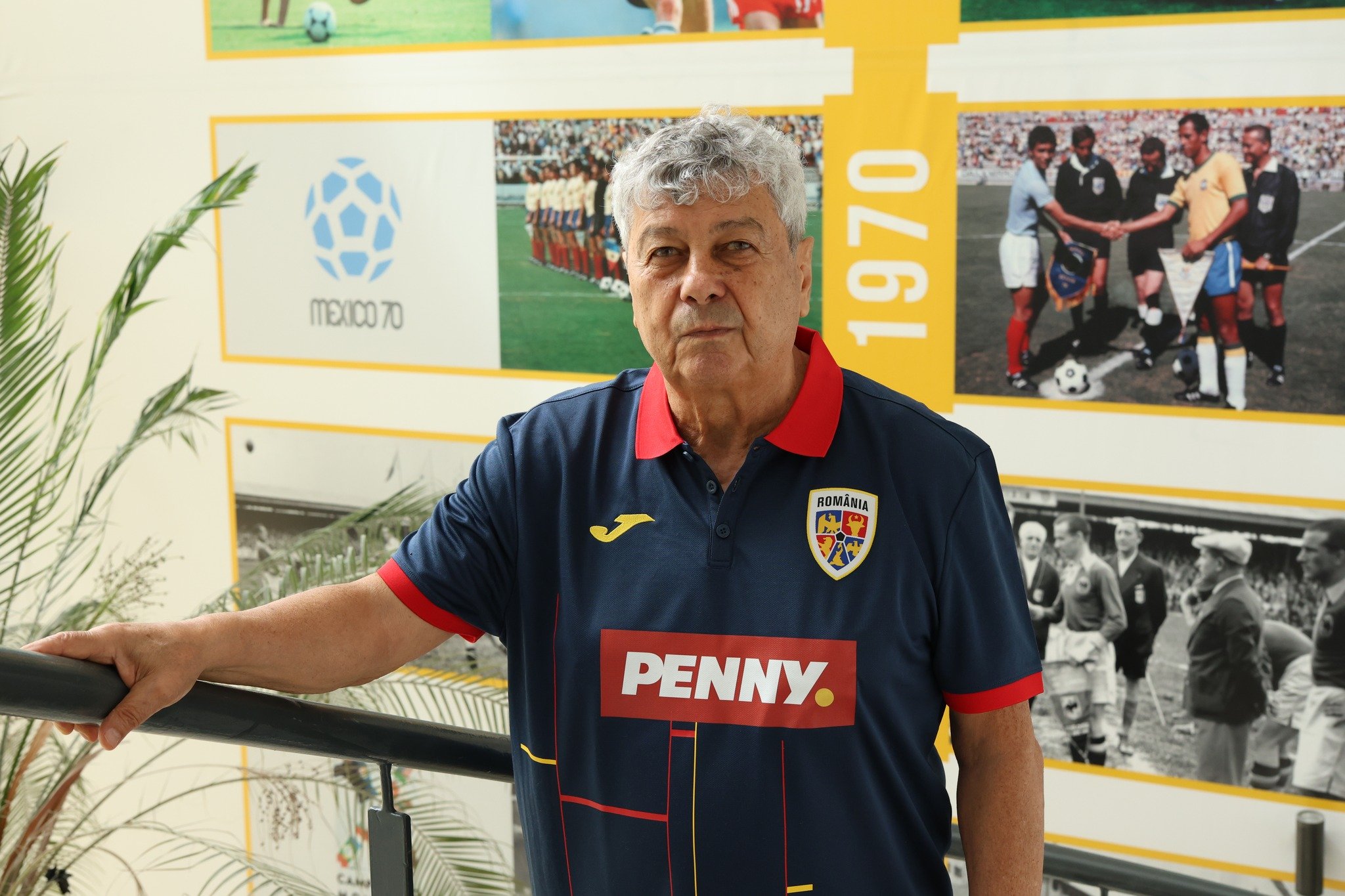 Mircea Lucescu sprijinit de o balustradă, &icirc;mbrăcat &icirc;n tricoul Naționalei, cu numele sponsorului Penny trecut mare pe piept și cu emblema echipei &icirc;n dreptul inimii