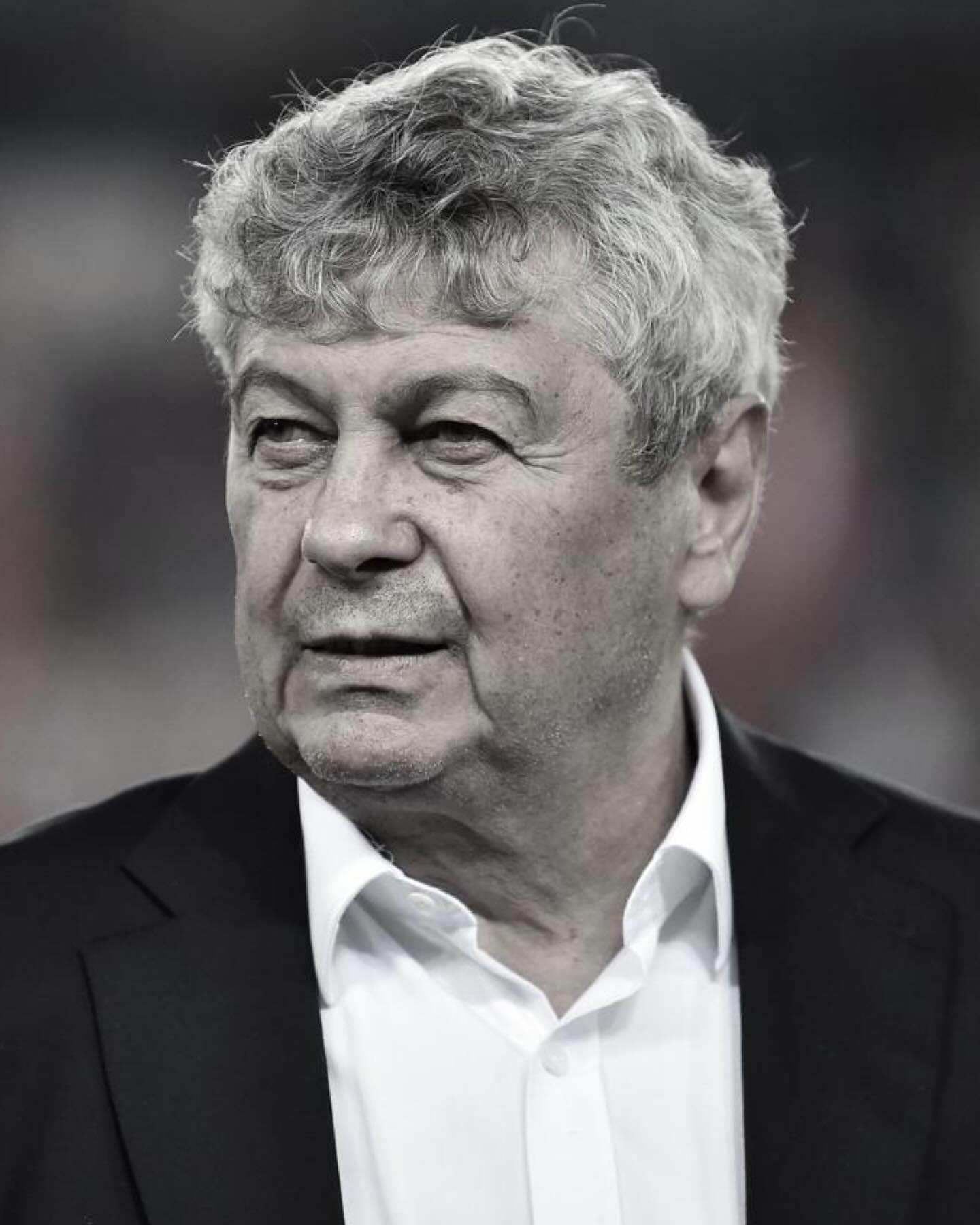 Imagine alb-negru cu Mircea Lucescu &icirc;n costum negru, cu cămașă albă, descheiată la g&acirc;t
