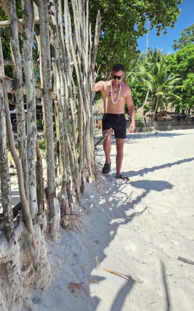 Valentin Sanfira la bustul gol, la plajă, &icirc;n Thailanda, purt&acirc;nd pantaloni scurți de culoare neagră, ochelari de soare și o ghirlandă cu flori la g&acirc;t