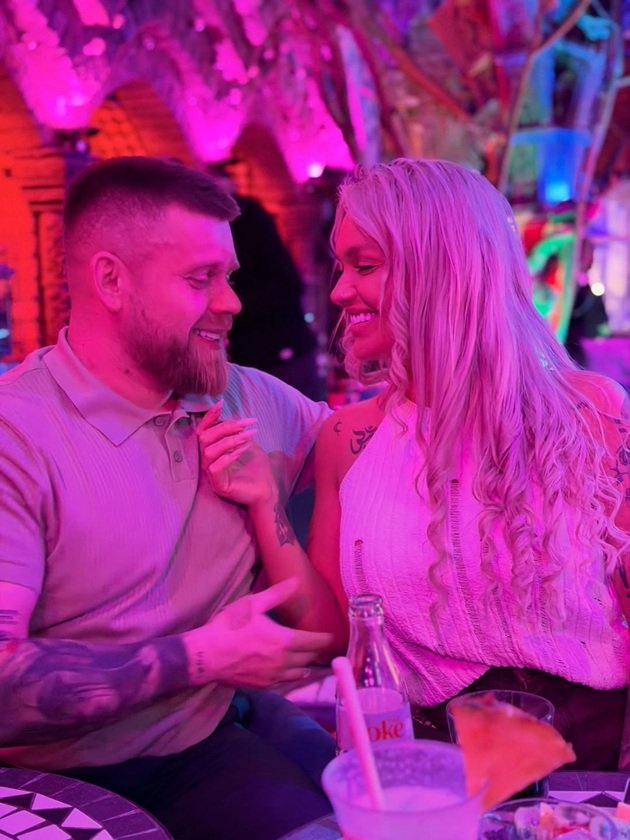 Răzvan Kovacs și Ella Vișan &icirc;ntr-un club, z&acirc;mbitori, st&acirc;nd la aceeași masă, &icirc;n timp ce femeia vrea să &icirc;l atingă subtil pe barbă