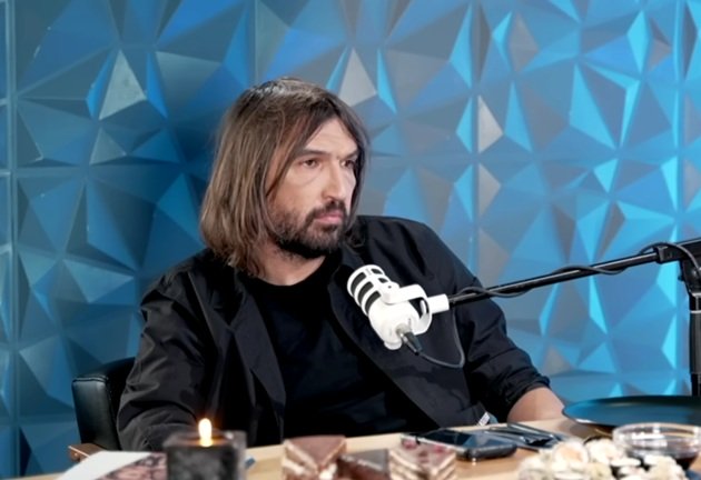 Dan Alexa st&acirc;nd pe scaun, la masă, față &icirc;n față cu Bursucu, &icirc;n timpul podcastului acestuia, av&acirc;nd &icirc;n față microfonul, sushi, prăjituri și o lum&acirc;nare aprinsă