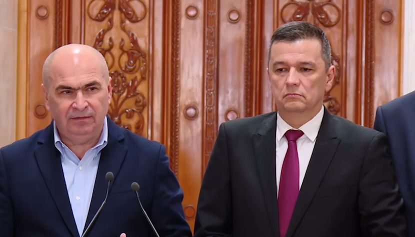 Ilie Bolojan și Sorin Grindeanu st&acirc;nd unul l&acirc;ngă altul &icirc;n cadrul unei &icirc;nt&acirc;lniri oficiale, &icirc;mbrăcați am&acirc;ndoi &icirc;n costum, st&acirc;nd la pupitru, &icirc;n fața unor microfoane