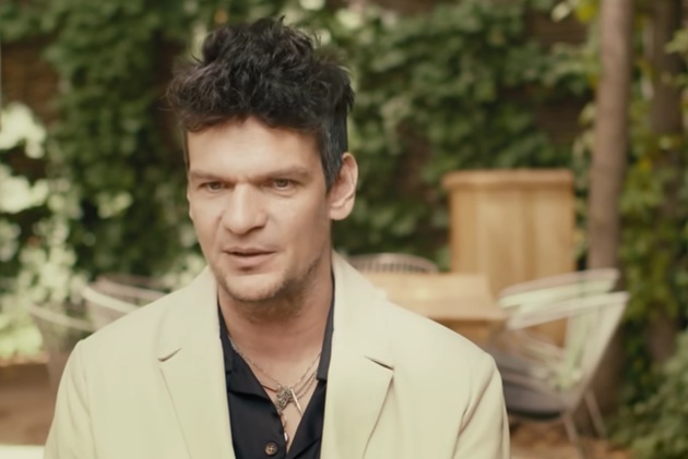 Tudor Chirilă &icirc;n cămașă neagră și costum bej, st&acirc;nd la masă, pe terasa unui restaurant, &icirc;n timpul filmării unui podcast