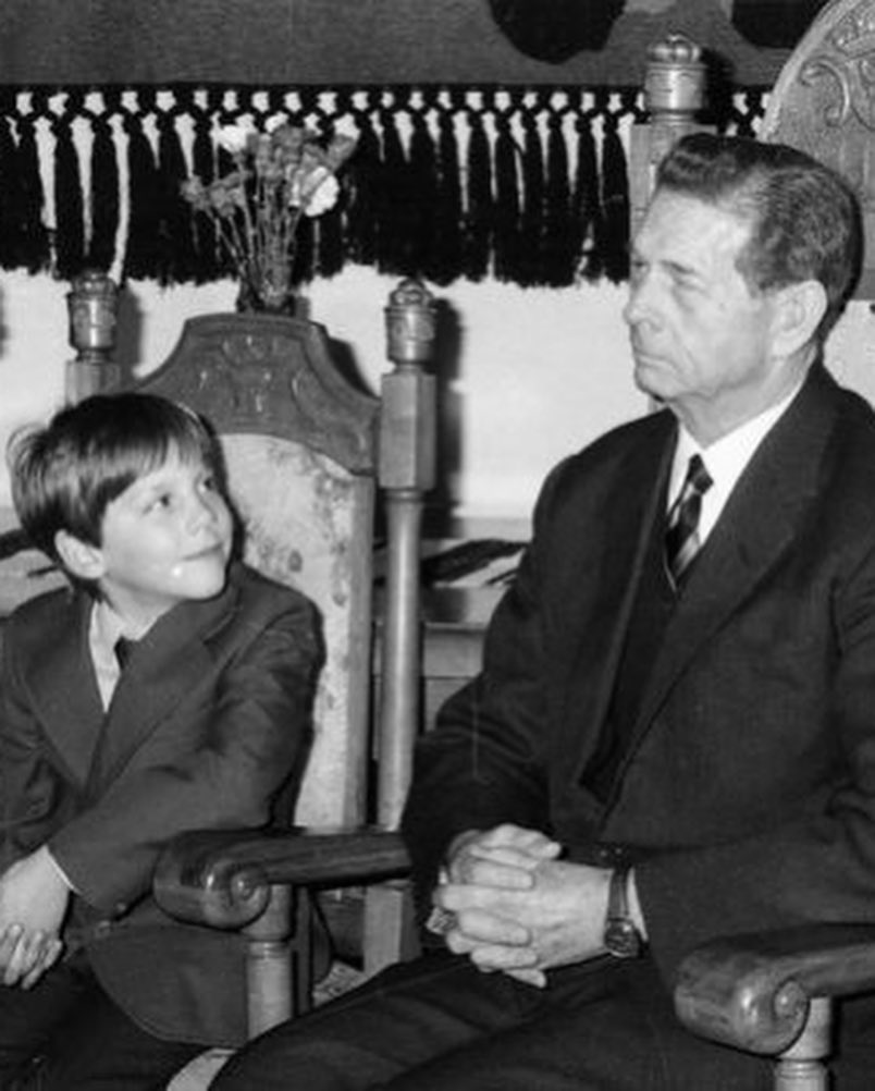 Imagine alb-negru cu Principele Nicolae &icirc;n copilărie, privindu-l cu admirație pe bunicul său, Regele Mihai, care stă l&acirc;ngă el pe un scaun, cu m&acirc;inile &icirc;ncrucișate, &icirc;n timp ce am&acirc;ndoi erau &icirc;mbrăcați elegant, &icirc;n costume &icirc;nchise la culoare