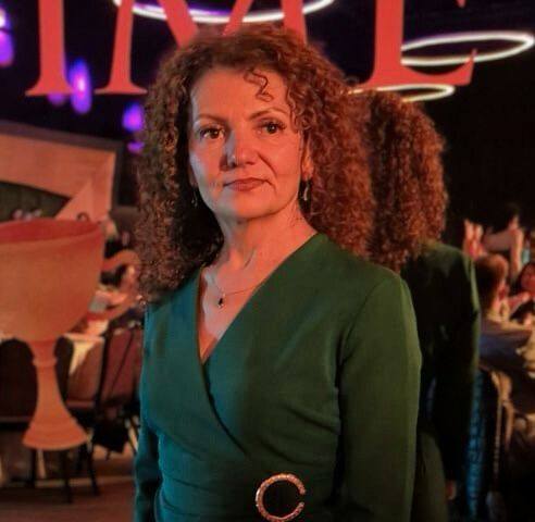 Florentina Bolojan cu părul creț și șaten, &icirc;mbrăcată &icirc;ntr-o rochie verde cu decolteu &icirc;n V și cataramă aurie &icirc;n talie