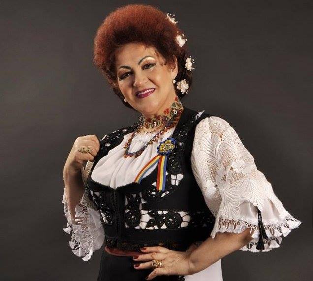 Elena&nbsp;Merișoreanu z&acirc;mbitoare, cu m&acirc;na &icirc;n șold, &icirc;mbrăcată &icirc;n costum popular, cu ie albă și vestă neagră brodată