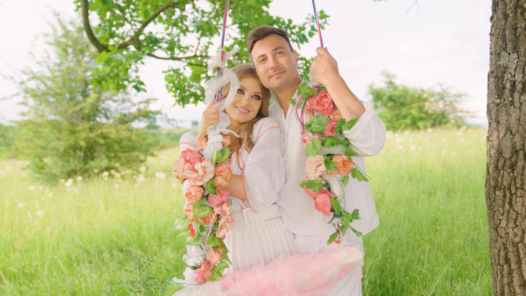 Valentin Sanfira și Codruța Filip st&acirc;nd &icirc;mpreună &icirc;n spatele unui leagăn din flori, undeva &icirc;n natură, l&acirc;ngă un copac, &icirc;n timp ce erau am&acirc;ndoi &icirc;mbrăcați &icirc;n alb