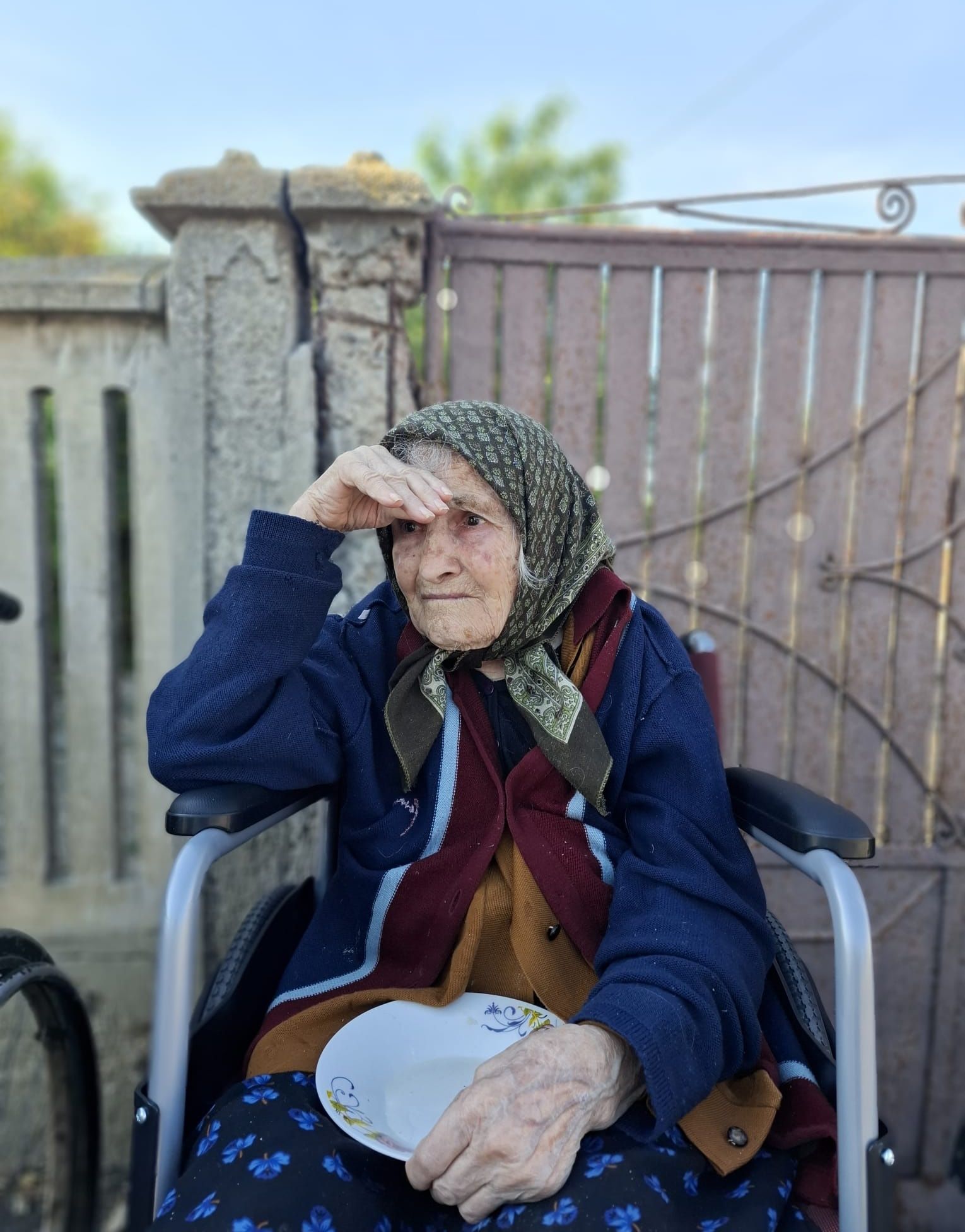 Bunica Oanei Lis, &icirc;n scaun cu rotile la poartă, &icirc;n timp ce ține &icirc;n m&acirc;nă o farfurie goală