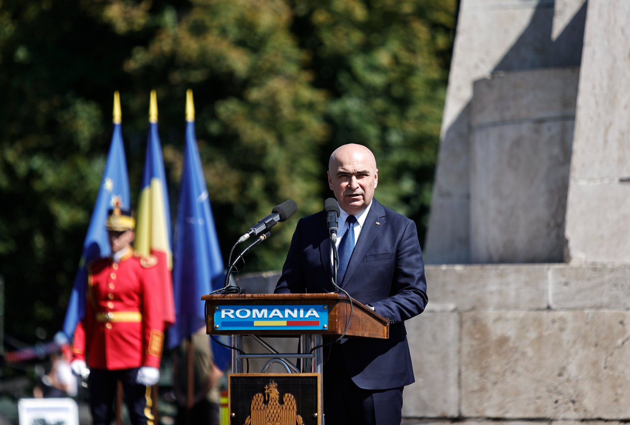 Ilie Bolojan vorbind la pupitru, &icirc;n fața oamenilor, la ceremonia dedicată Zilei Aviației și a Forțelor Aeriene, &icirc;mbrăcat &icirc;ntr-un costum bleumarin cu cămașă albă și cravată albastră