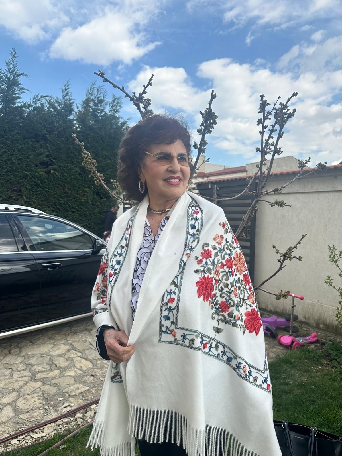 Irina Loghin st&acirc;nd &icirc;n curte, l&acirc;ngă un copac &icirc;nmugurit, &icirc;mbrăcată &icirc;ntr-o bluză albă cu volane cu margini negre, purt&acirc;nd pe umeri un șal alb cu imprimeu floral &icirc;n nuanțe de roșu, portocaliu și verde