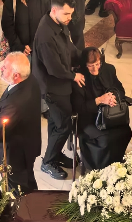 Mama lui Ioan Isaiu îmbrăcată în negru, stând pe scaun lângă sicriul actorului, la priveghi, în timp ce era sprijinită de un tânăr din familie
