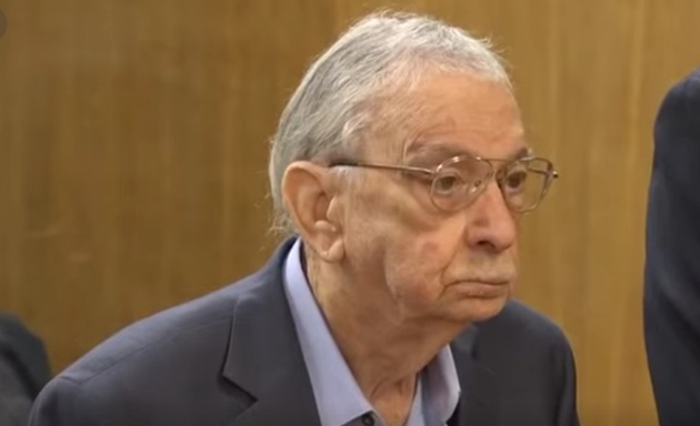 John Bernard Feit la peste 80 de ani, st&acirc;nd &icirc;n fața judecătorilor c&acirc;nd a fost condamnat la &icirc;nchisoare, &icirc;mbrăcat &icirc;n costum gri &icirc;nchis și cămașă bleu