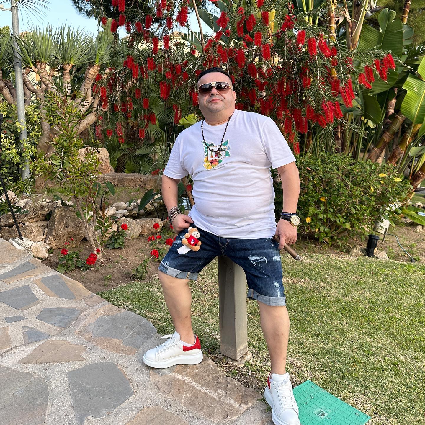 Adrian Minune st&acirc;nd relaxat &icirc;n fața camerei de fotografiat, &icirc;ntr-un tricou alb și o pereche de pantaloni scurți de blugi, țin&acirc;nd &icirc;n m&acirc;nă un trabuc