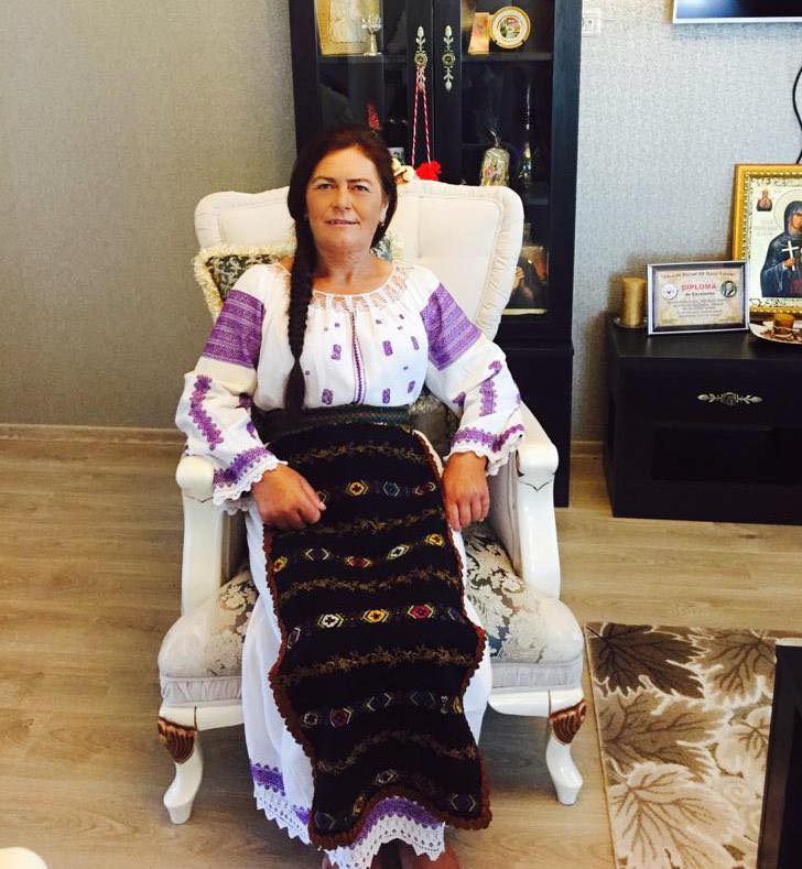 Elena, mama lui Valentin Sanfira, st&acirc;nd pe un fotoliu, &icirc;mbrăcată &icirc;n costum popular, cu ie albă cu model mov și fote negre cu modele colorate