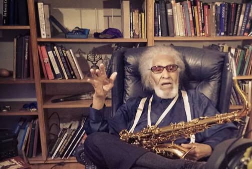 Sonny Rollins &icirc;mbrăcat &icirc;n pantaloni negri, cămașă bleumarin, st&acirc;nd pe un fotoliu de piele, cu saxofonul &icirc;n brațe