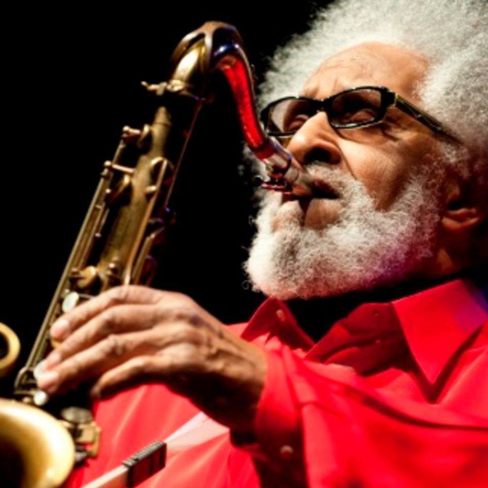 Sonny Rollins cu părul creț și alb, &icirc;mbrăcat &icirc;n costum roșu, pe scenă, c&acirc;nt&acirc;nd la saxofon