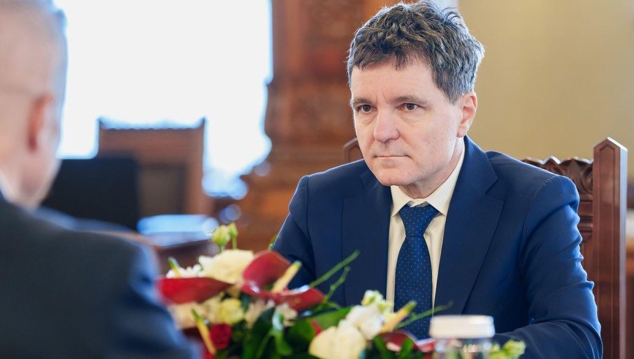 Președintele Nicușor Dan, st&acirc;nd serios la un birou, &icirc;mbrăcat &icirc;n costum albastru, cămașă albă și cravată albastră cu picățele