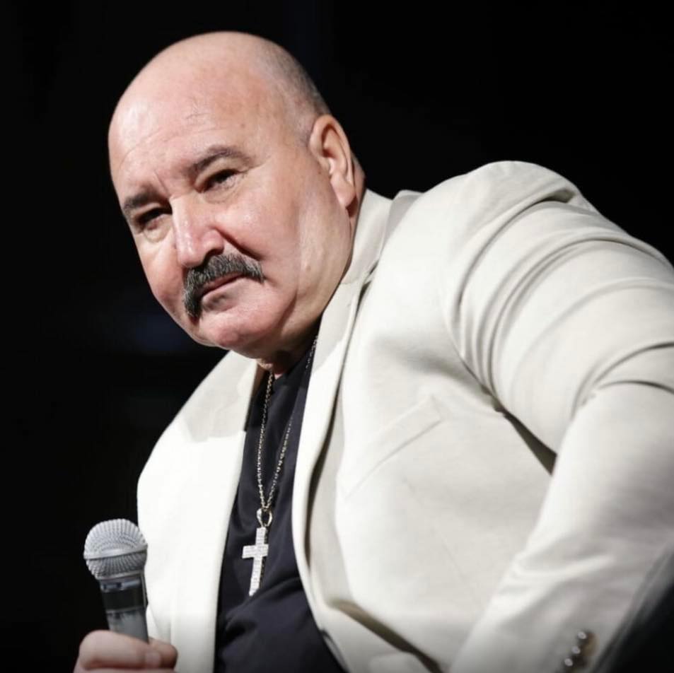 Nuțu Cămătaru cu microfonul &icirc;n m&acirc;nă, &icirc;ntr-un tricou negru și un sacou gri deschis, purt&acirc;nd o cruce de mari dimensiuni la g&acirc;t