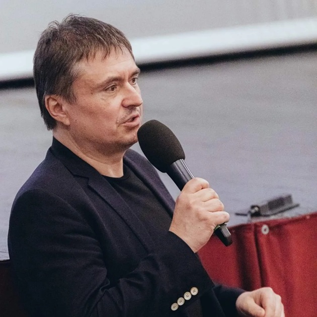 Regizorul Cristian Mungiu, la un eveniment cultural, vorbind la microfon &icirc;n fața publicului, &icirc;n timp ce era &icirc;mbrăcat &icirc;n tricou negru și sacou de aceeași culoare
