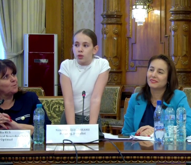 Olteanu Ilinca Maria, &icirc;n fața microfonului, &icirc;ntr-un tricou alb, vorbind &icirc;n Parlament, &icirc;n fața tuturor celor invitați la dezbaterea despre sănătatea mintală a copiilor