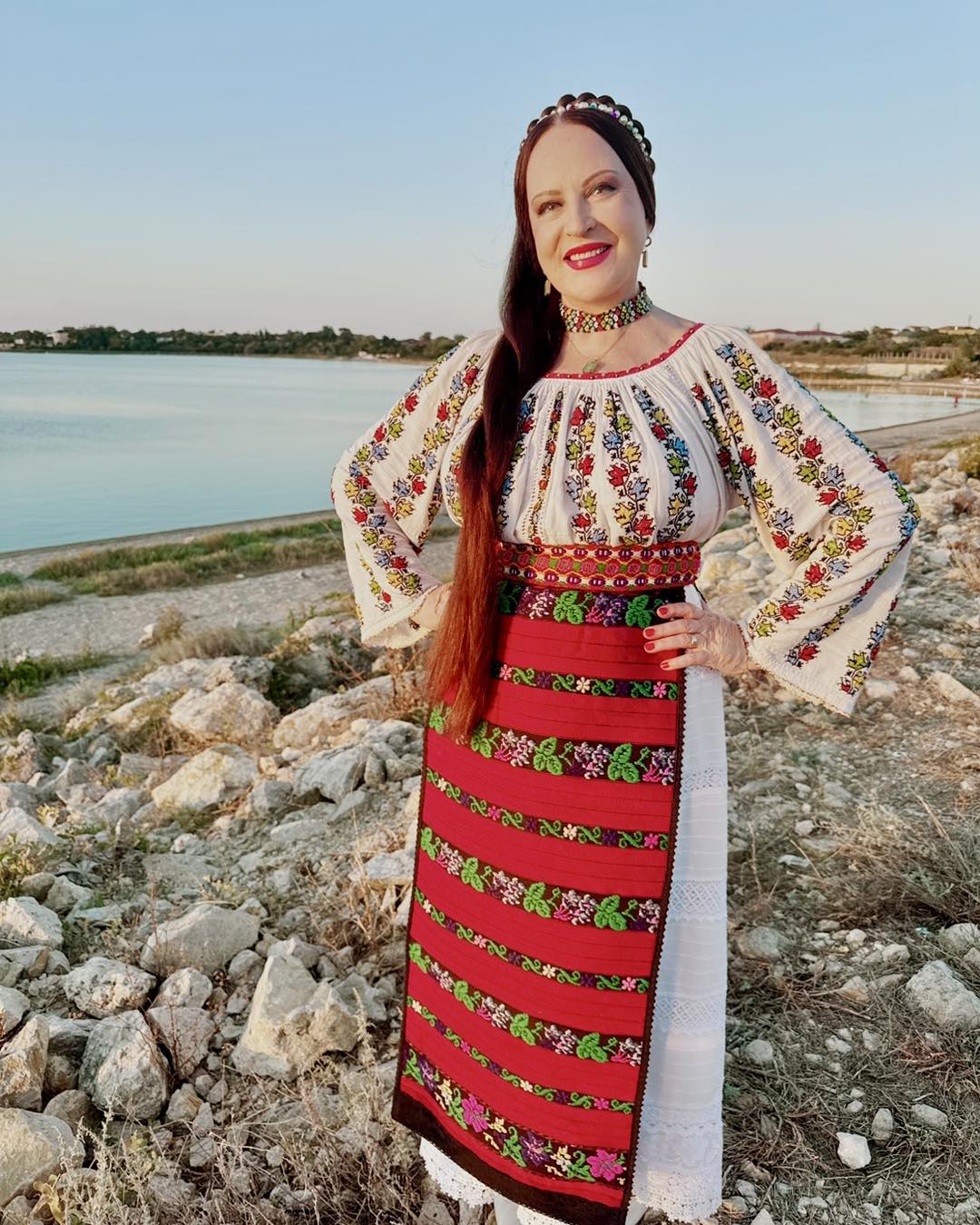 Maria Dragomiroiu în costum popular, cu părul despletit și dat într-o parte, stând cu mâinile în șold pe malul unui lac