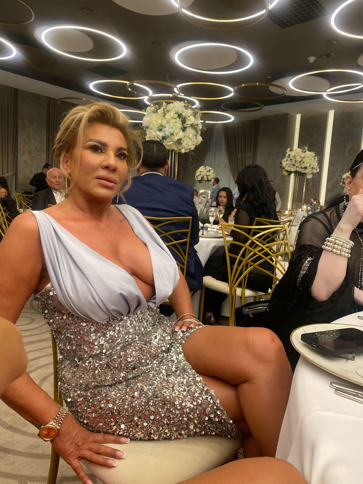 Lidia Balint, prima soție a lui Nuțu Cămătaru, &icirc;mbrăcată &icirc;ntr-o rochie gri, cu paiete argintii, st&acirc;nd pe scaun, la masa unui restaurant, &icirc;n timpul unui eveniment
