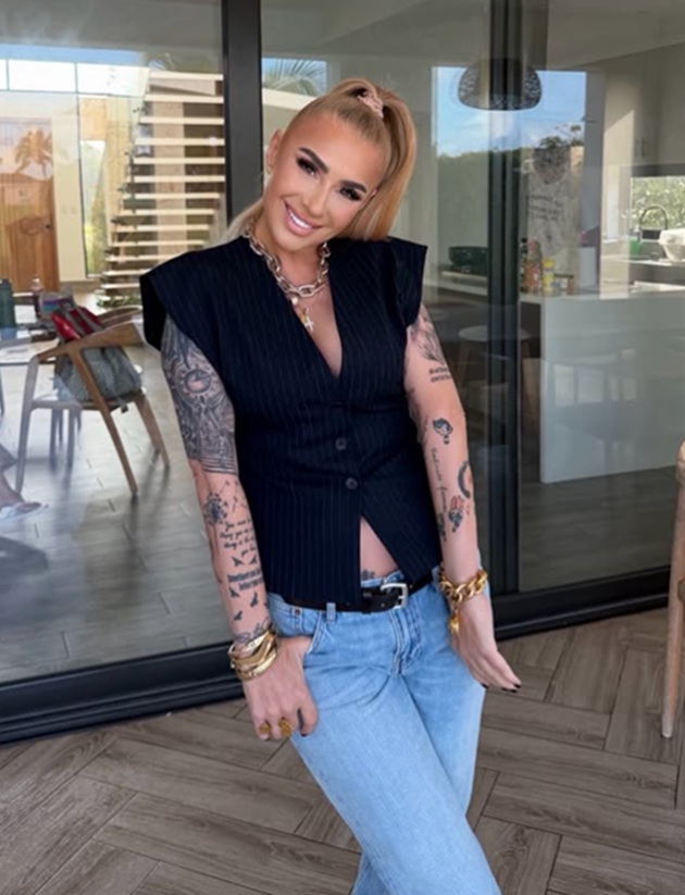 Anamaria Prodan zâmbitoare, în blugi deschiși la culoare și în top negru cu dungi, ținându-și degetele mari în buzunarele de la blugi, în timp ce purta bijuterii masive
