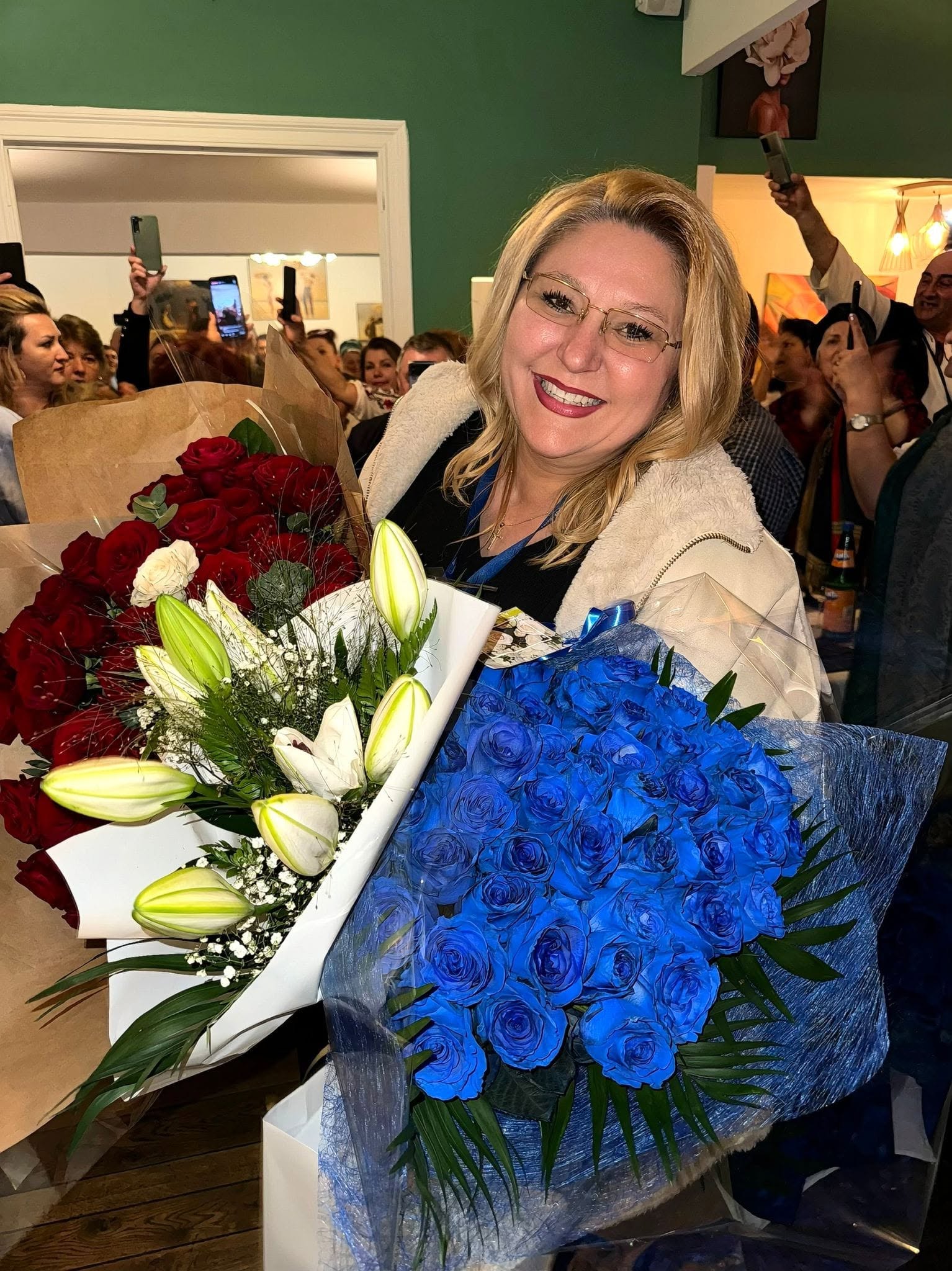 Diana Șoșoacă z&acirc;mbitoare, &icirc;mbrăcată &icirc;n negru, cu o jachetă bej, țin&acirc;nd &icirc;n brațe mai multe buchete de flori